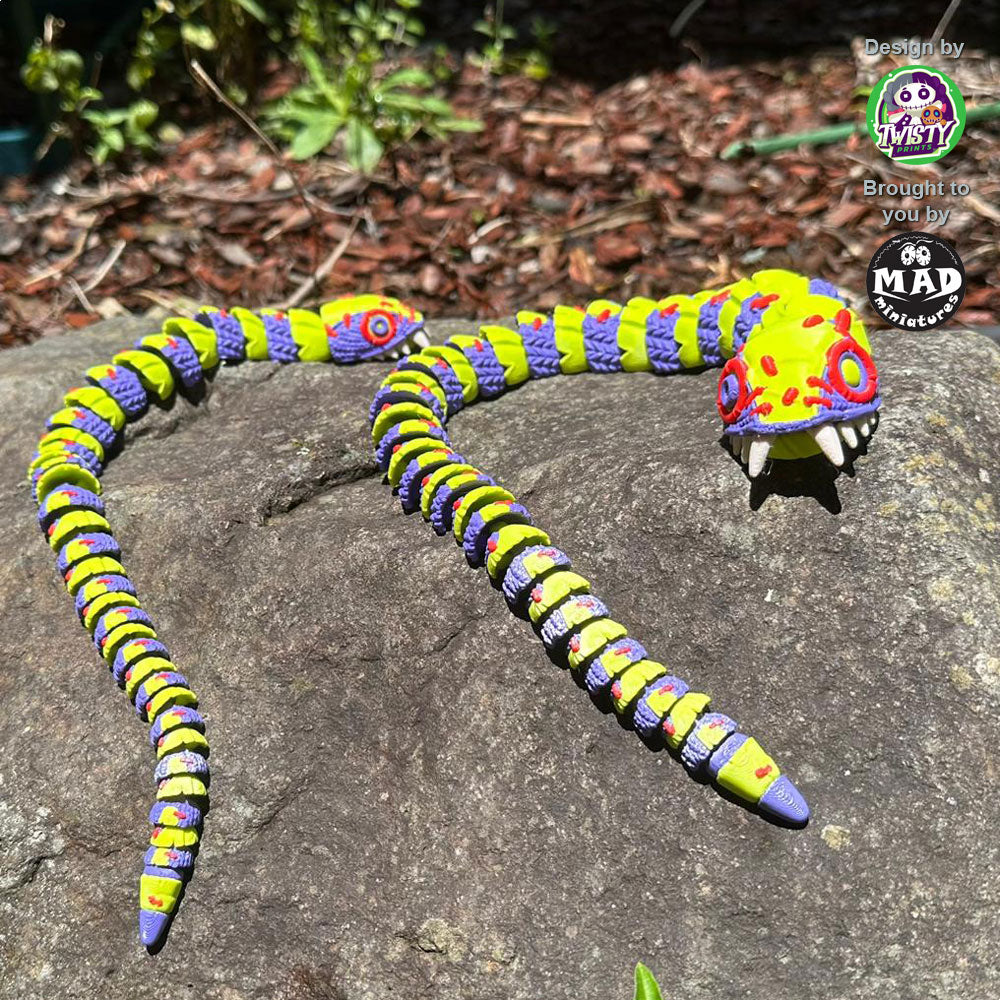 Stuffed Snake (both sizes) sunbathing on a rock, showcasing their vibrant yellow-green and purple patchwork design, with mismatched eyes and bold red stitches.