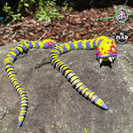 Stuffed Snake (both sizes) sunbathing on a rock, showcasing their vibrant yellow-green and purple patchwork design, with mismatched eyes and bold red stitches.