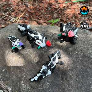 Cute Sweetheart Skunks from Mad Miniatures, designed by Cinderwing, on a rock, showcasing red, pink, and purple rose variations and one without a rose. Perfect for collectors.